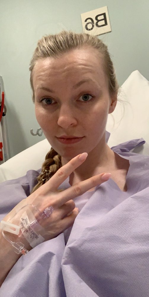 Heather in a hospital bed before surgery, peace sign